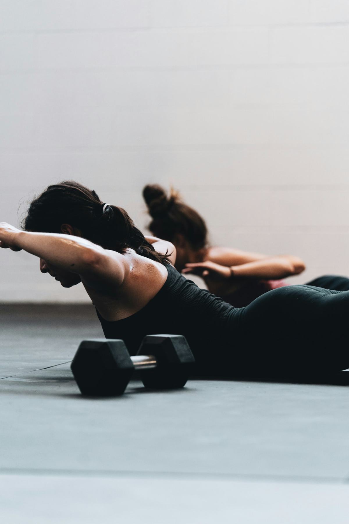 Two women lying face-down on a fitness studio floor, lifting their chests and bent arms off the ground in a back extension exercise, a black hex dumbbell resting on the floor in front of them.