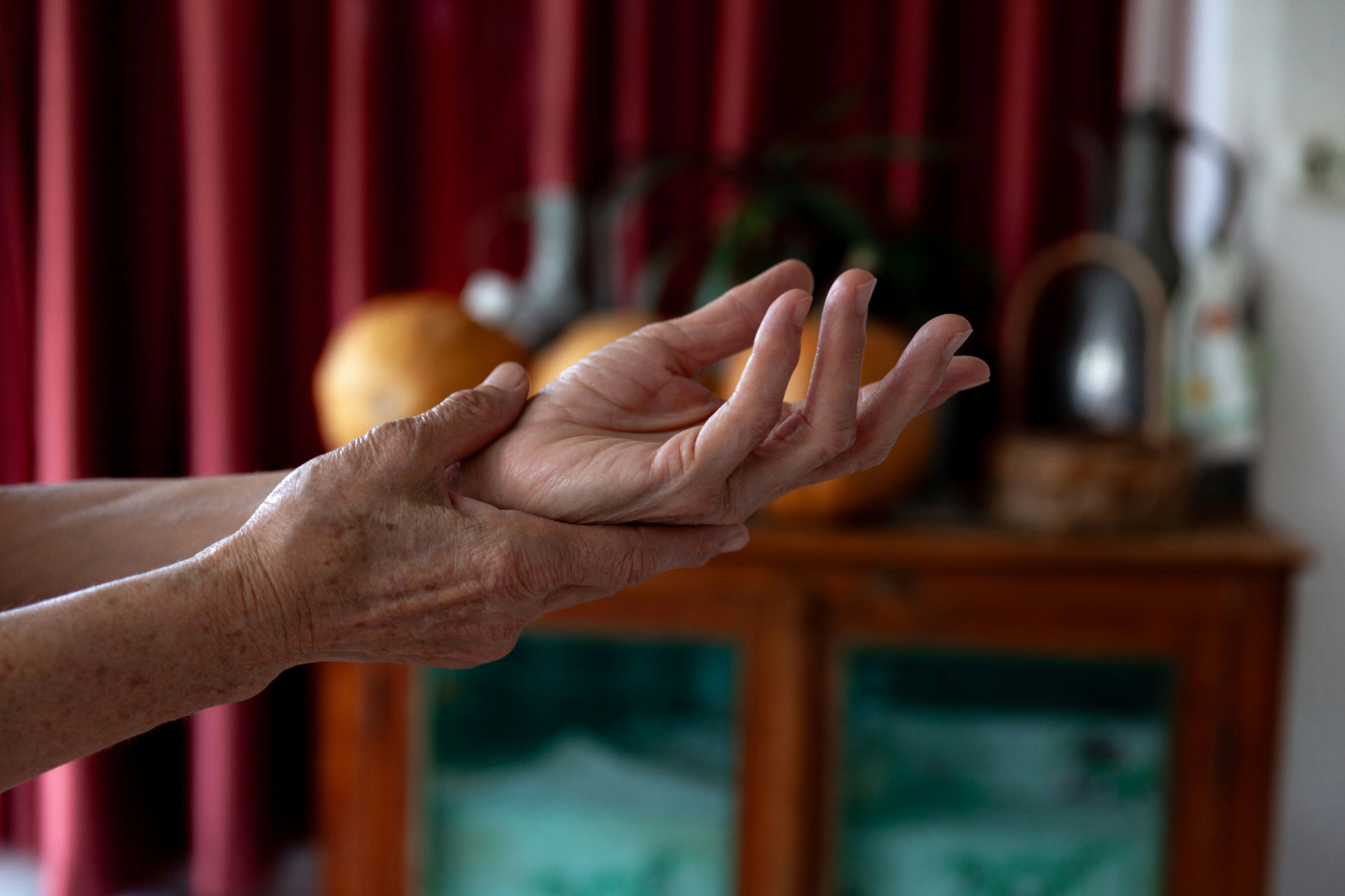 An older adult's hands in a warm-lit room, one hand gently supporting the other wrist, soft red drapery and a wooden cabinet behind.